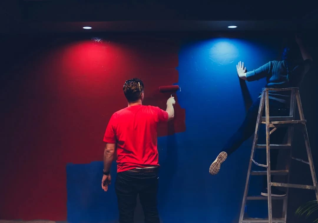 Two people painting a wall, one in a red t-shirt with a roller, another on a ladder, wall half red half blue