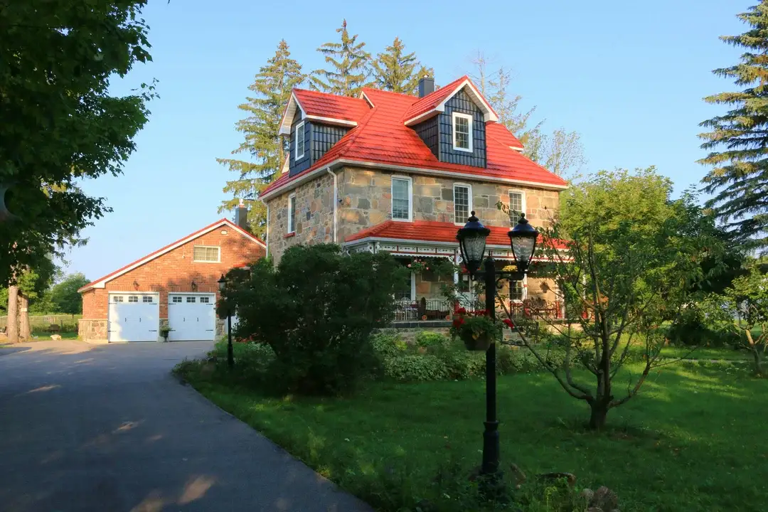 Maison en pierre avec toiture en métal rouge, galerie enveloppante et jardin fleuri, architecture patrimoniale québécoise