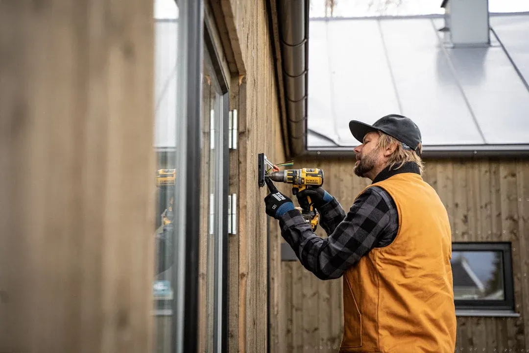 Technicien en veste orange avec perceuse DeWalt fixant une fenêtre sur une façade en bois