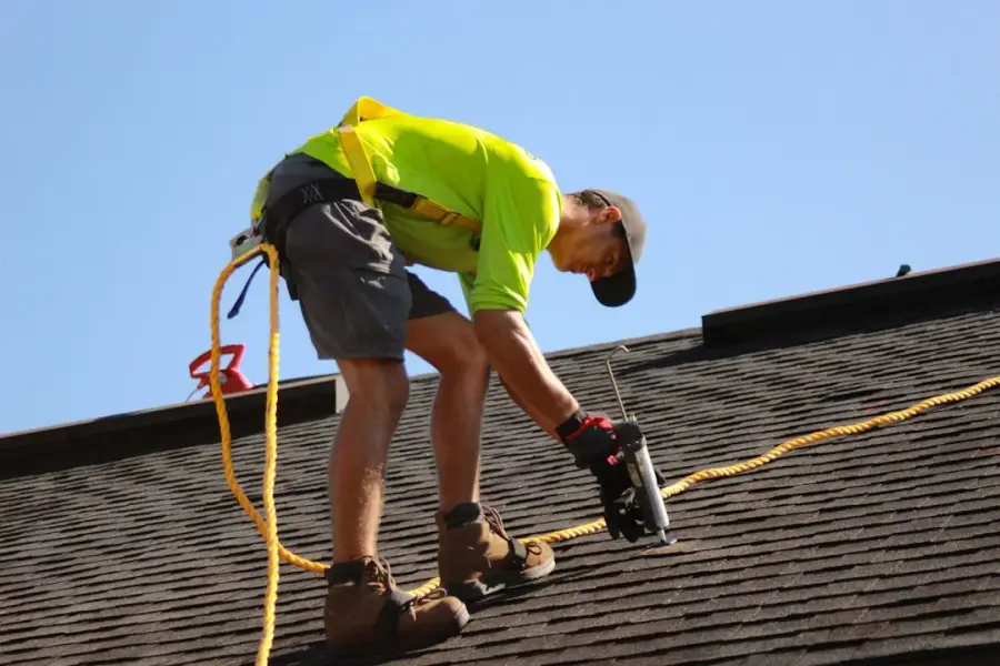 Roofer in yellow hi-vis vest with safety harness and caulk gun working on dark asphalt shingles against blue sky
