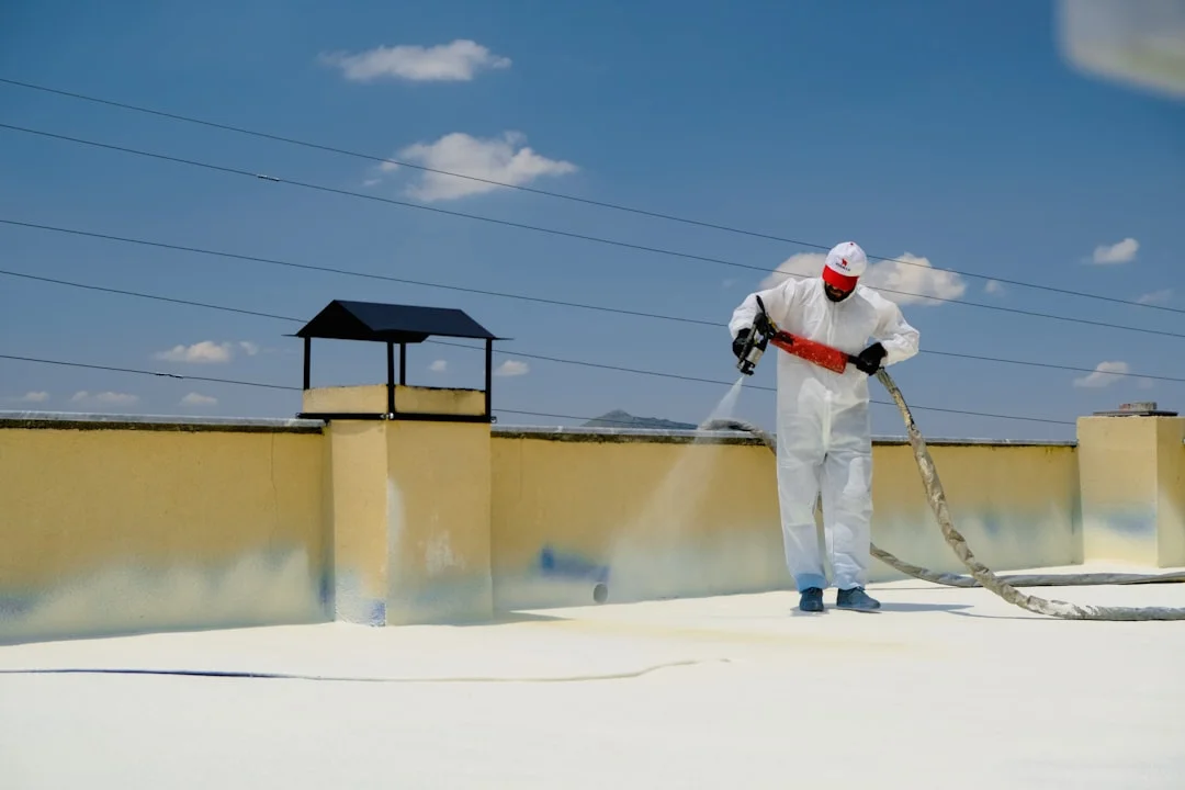 Technicien en combinaison blanche appliquant de la mousse isolante avec un pistolet sur un toit plat sous ciel bleu
