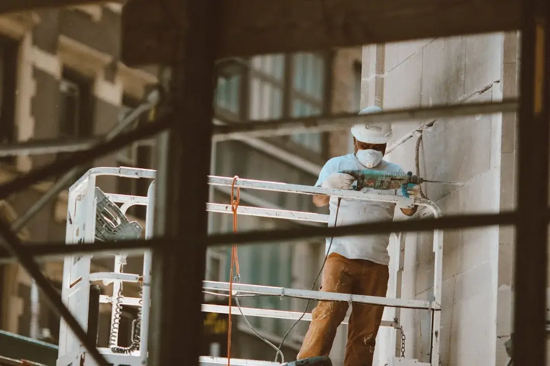 Ouvrier en casque blanc et masque sur échafaudage métallique utilisant un marteau perforateur sur un mur de pierre